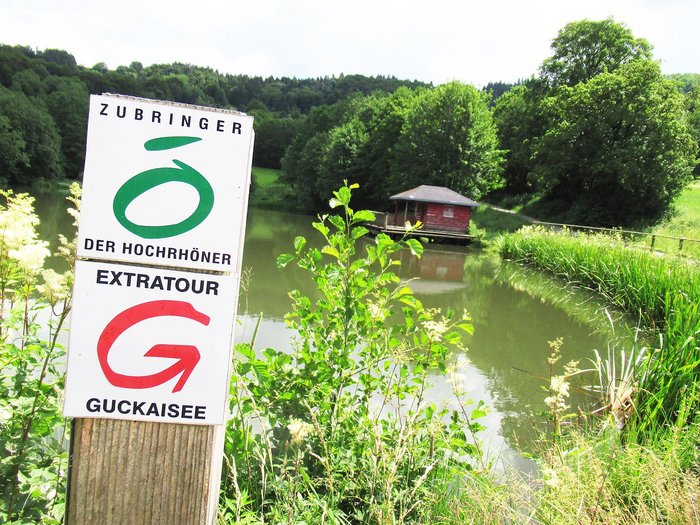Markierung der Gucksaisee-Tour vor einem Gewässer mit Hütte im Hintegrund